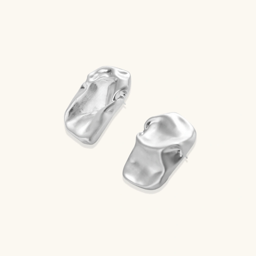 Pair of sculptural silver ear cuffs with organic fluid shapes and polished finish against cream background.