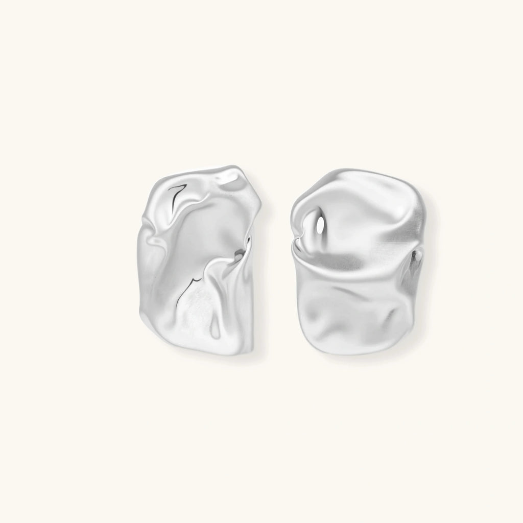 Pair of sculptural silver ear cuffs with organic fluid shapes and polished finish against cream background.