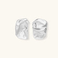 Pair of sculptural silver ear cuffs with organic fluid shapes and polished finish against cream background.